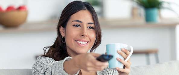 woman smiling and drinking a hot beverage while watching tv